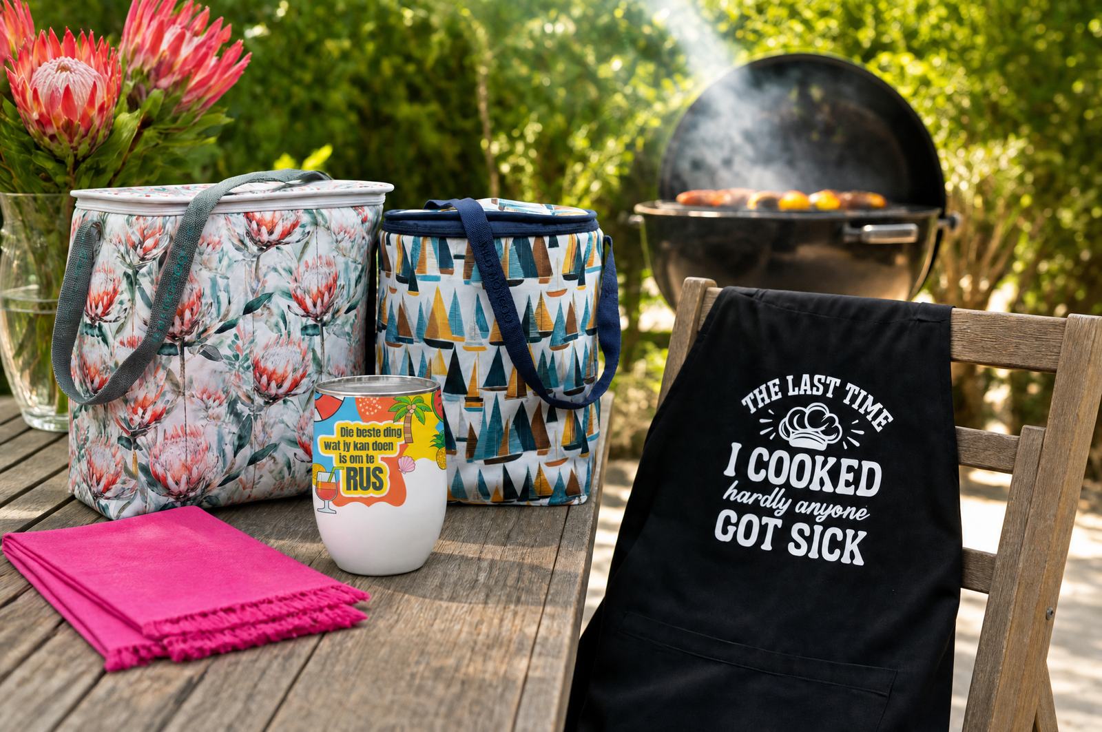 Wild Mongoo protea cooler bag, sailboat cooler, RUS gin tumbler and funny braai apron on a garden table in George