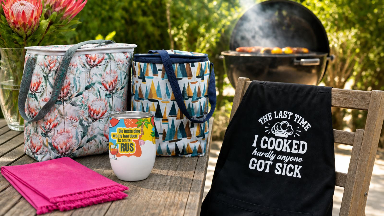 Wild Mongoo protea cooler bag, sailboat cooler, RUS gin tumbler and funny braai apron on a garden table in George