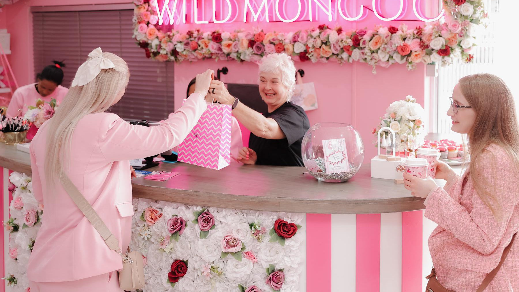 A customer being handed a pink Wild Mongoo gift bag across the flower-covered counter in the George shop, under the neon sign